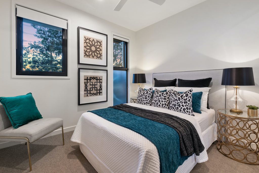 An image of a bedroom showing a large double bed in the centre, with blue dressing, a bedside table with lamp, large windows to the left with some art in between, finally a seat in the corner of the room.